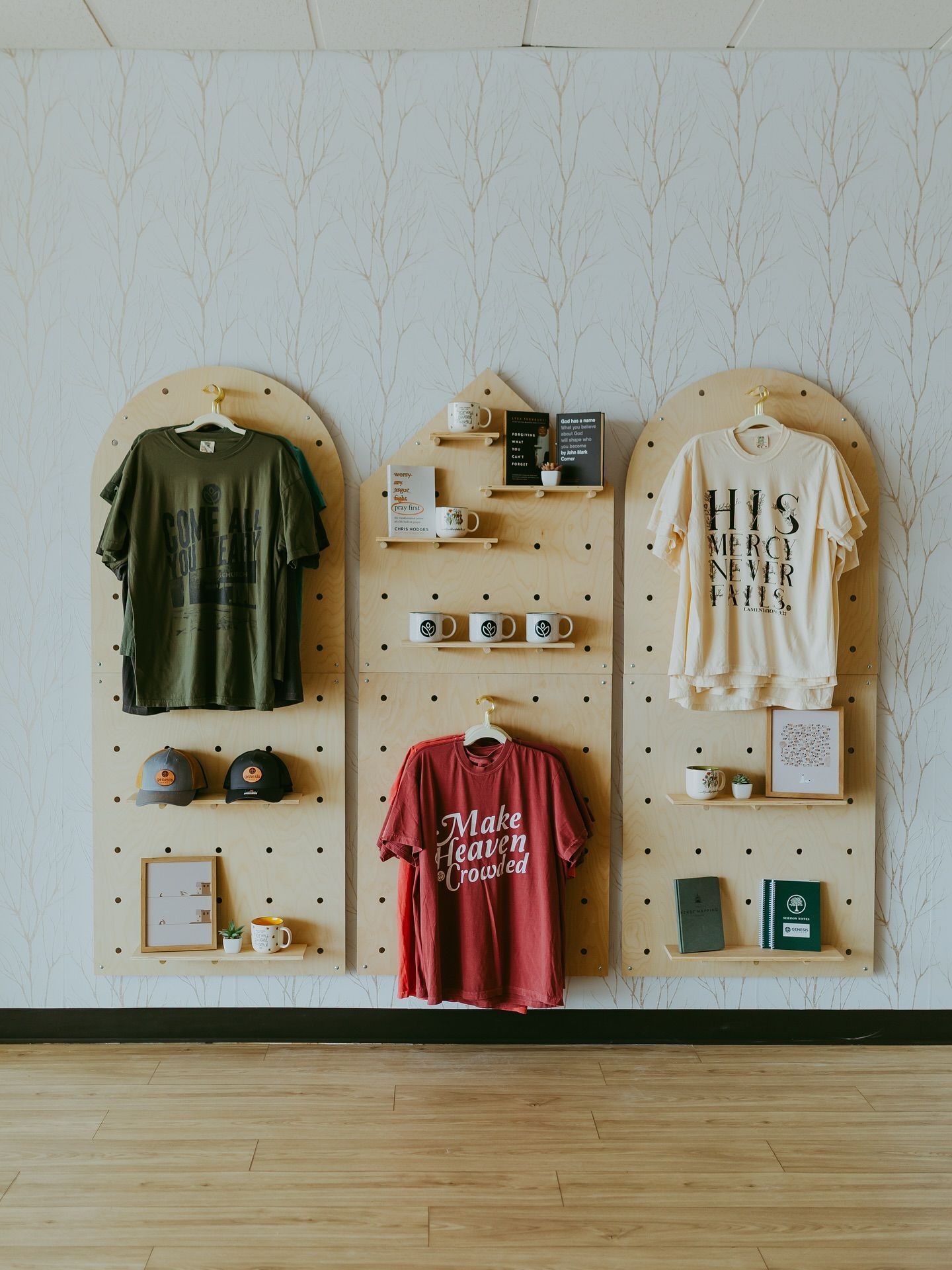Retail display with three wooden pegboards showcasing shirts, mugs, hats, and small items against a white wall.