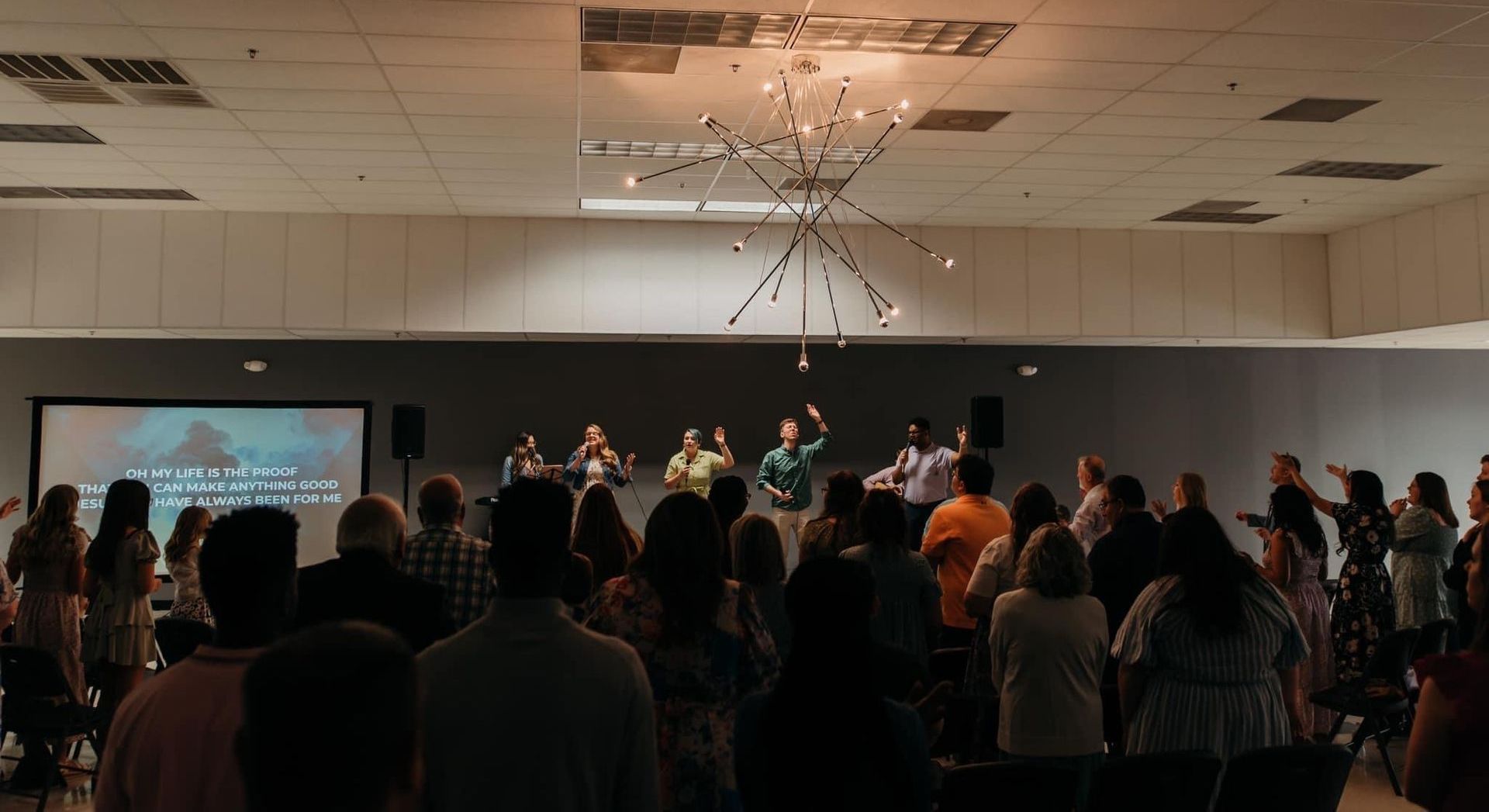 Worship service in a large room. People raise their hands toward a stage with a band and screen displaying lyrics.