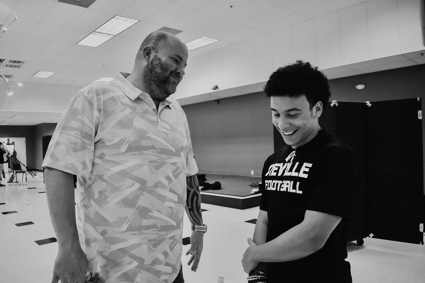 Two men smiling and talking indoors; one wears a patterned shirt, the other a 