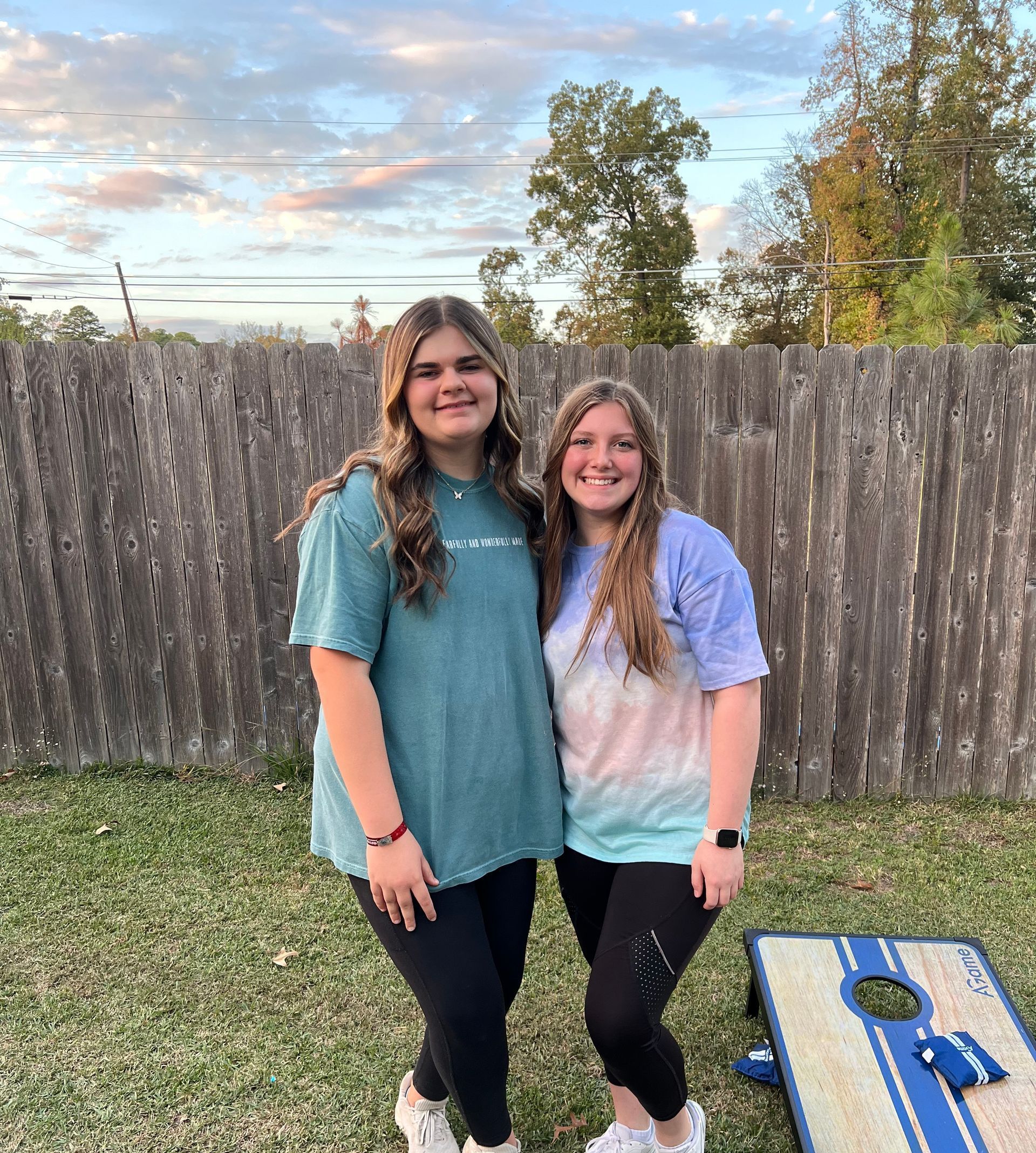 Two young women are standing next to each other in front of a wooden fence.
