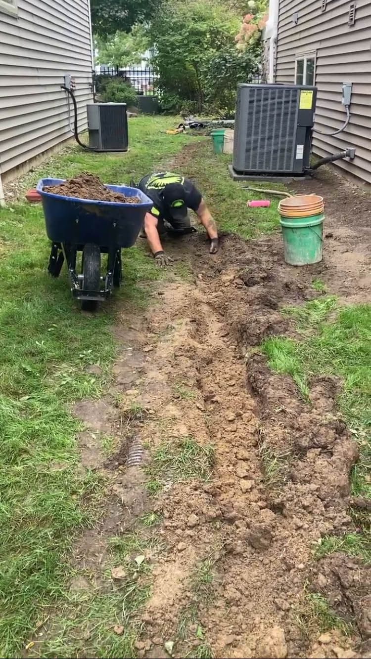 A man is digging in the dirt in a yard with a wheelbarrow.