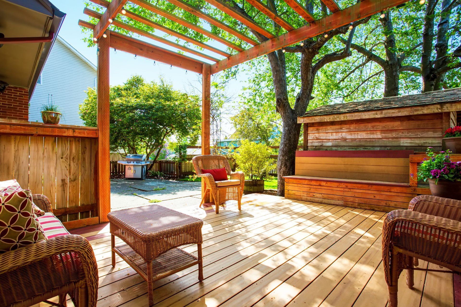A cozy backyard deck in Chula Vista, CA, bathed in sunlight, features natural wood planks and a pergola with an open beam design. Rattan furniture with cushioned seats invites relaxation, while a built-in hot tub promises leisure. Lush greenery, including a mature tree, peeks over a high wooden fence, creating a private outdoor retreat. String lights dangle above, suggesting a warm ambiance for evenings, and vibrant potted flowers add a splash of color to the tranquil setting.