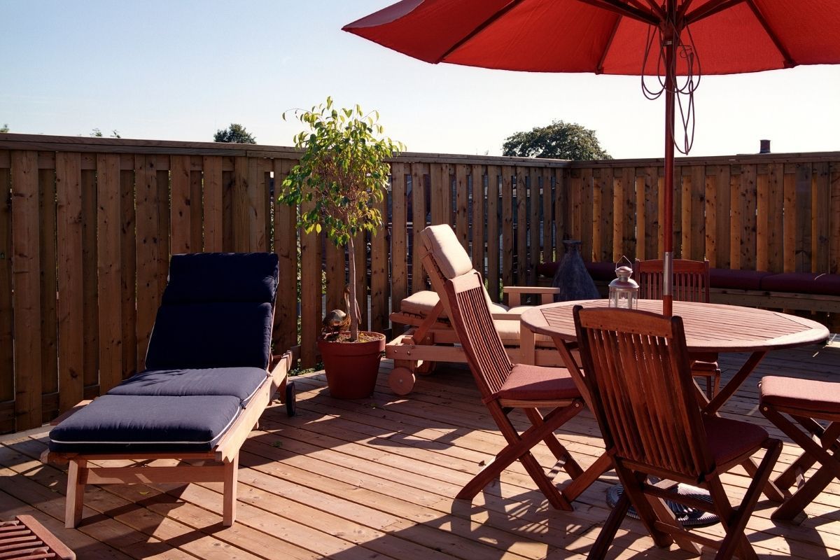 A cozy deck space in Chula Vista, CA, featuring wooden furniture including loungers with navy cushions, a round table, and folding chairs. A vibrant red umbrella offers a pleasant shade on a sunny day. Potted plants add a touch of greenery, enhancing the deck’s welcoming vibe. The wooden fence around the deck ensures privacy, creating an intimate setting for relaxation or social gatherings. The scene is bathed in sunlight, suggesting a warm, inviting outdoor atmosphere.