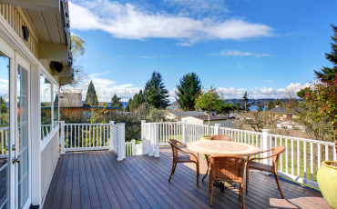 A photo of a newly built deck. The deck has a white railing surrounding the deck. The deck is installed on the second level of he house. There is a staircase towards the back of the photo that will lead you from the deck down to the grass.