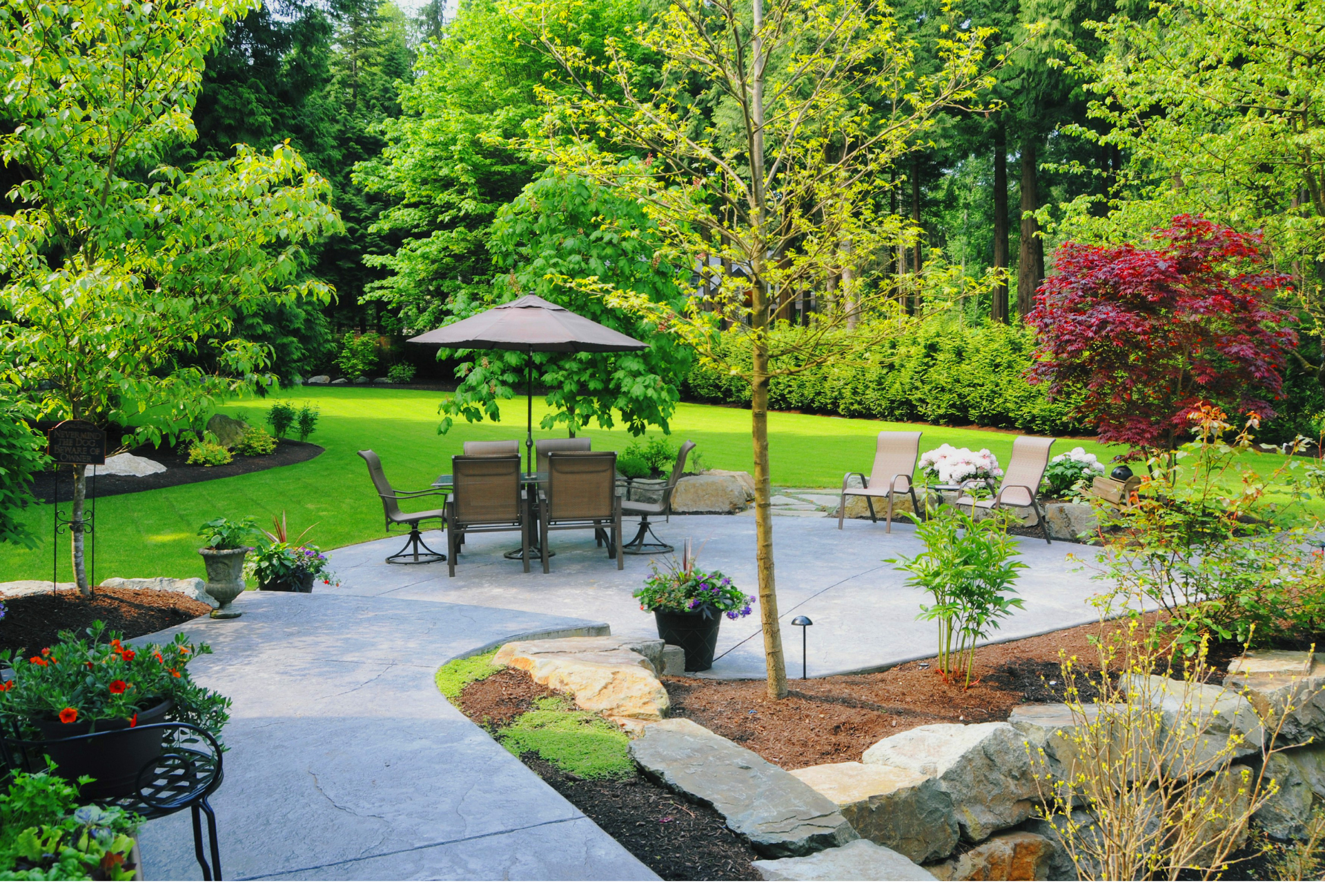 A beautiful photo of the backyard of a home. The backyard has bright green grass and a wall of trees in the distance. There is a patio in the middle of the photo. The patio has beautiful flowers and plants surrounding its perimeter. The patio also has lawn furniture to welcome guests.