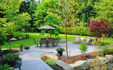 A photo of a patio that is in the backyard of a chula vista home. The Patio has beautiful landscaping surrounding it with multiple trees, plants, and flowers. The patio has nice patio furniture sitting at the end.