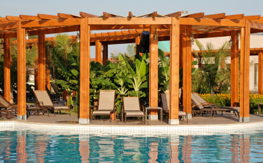 A photo of a pergola in a luxury home. The pergola is a substantial size. The pergola was built right next to a pool, so there are pool chairs along with plants sitting underneath the pergola. This is a beautiful photo of the work that South Bay Deck Pros can accomplish.