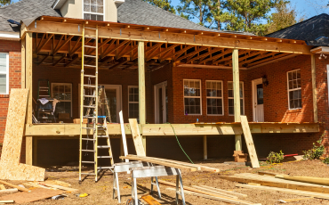 A photo of a house that is in the process of adding a new porch to their house. The Old porch was removed and this new one is being built. It is almost done being built. All that is left is railings, steps, and a coat of paint.