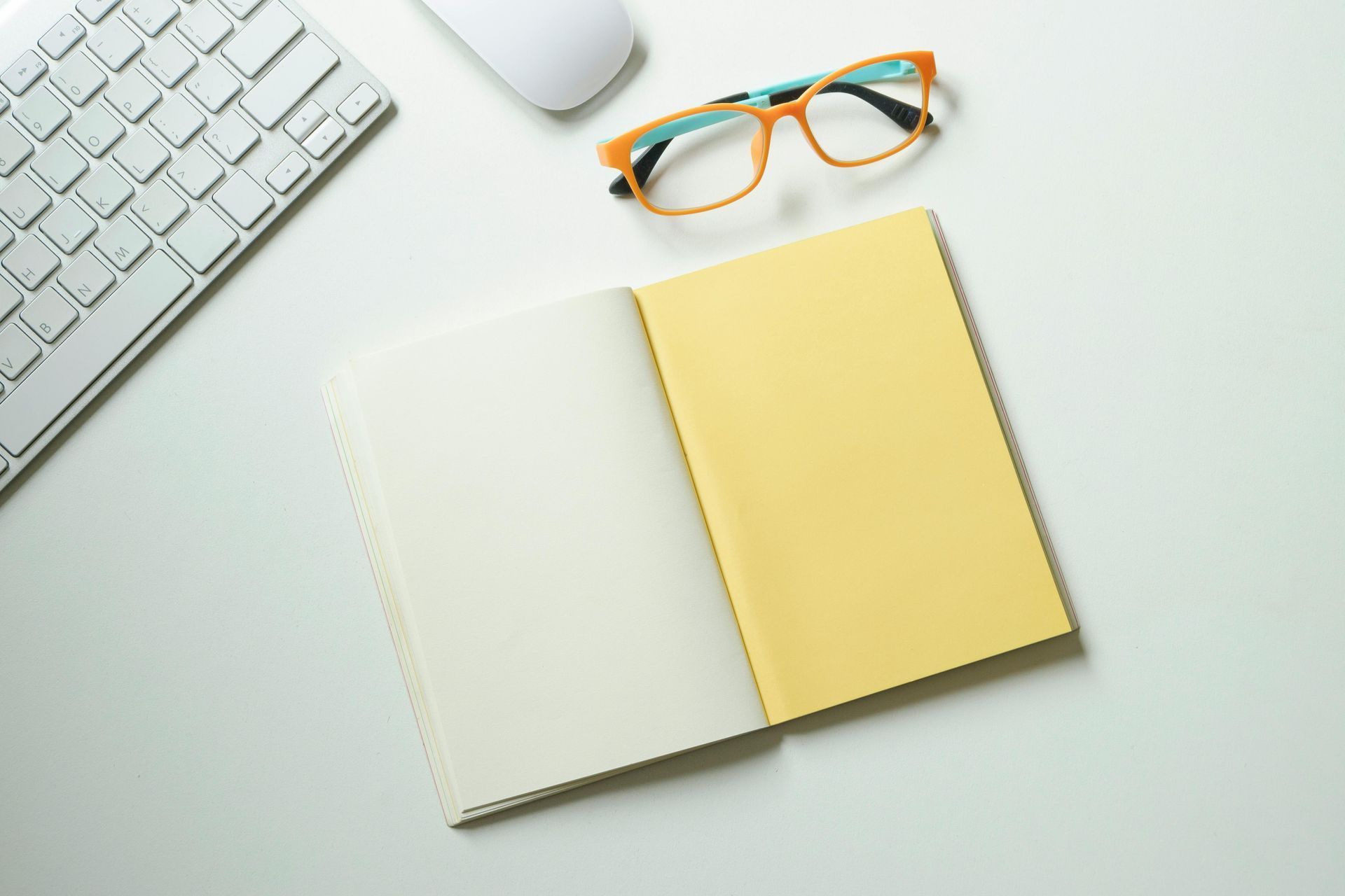 Open notebook with yellow cover beside glasses and keyboard on a white desk