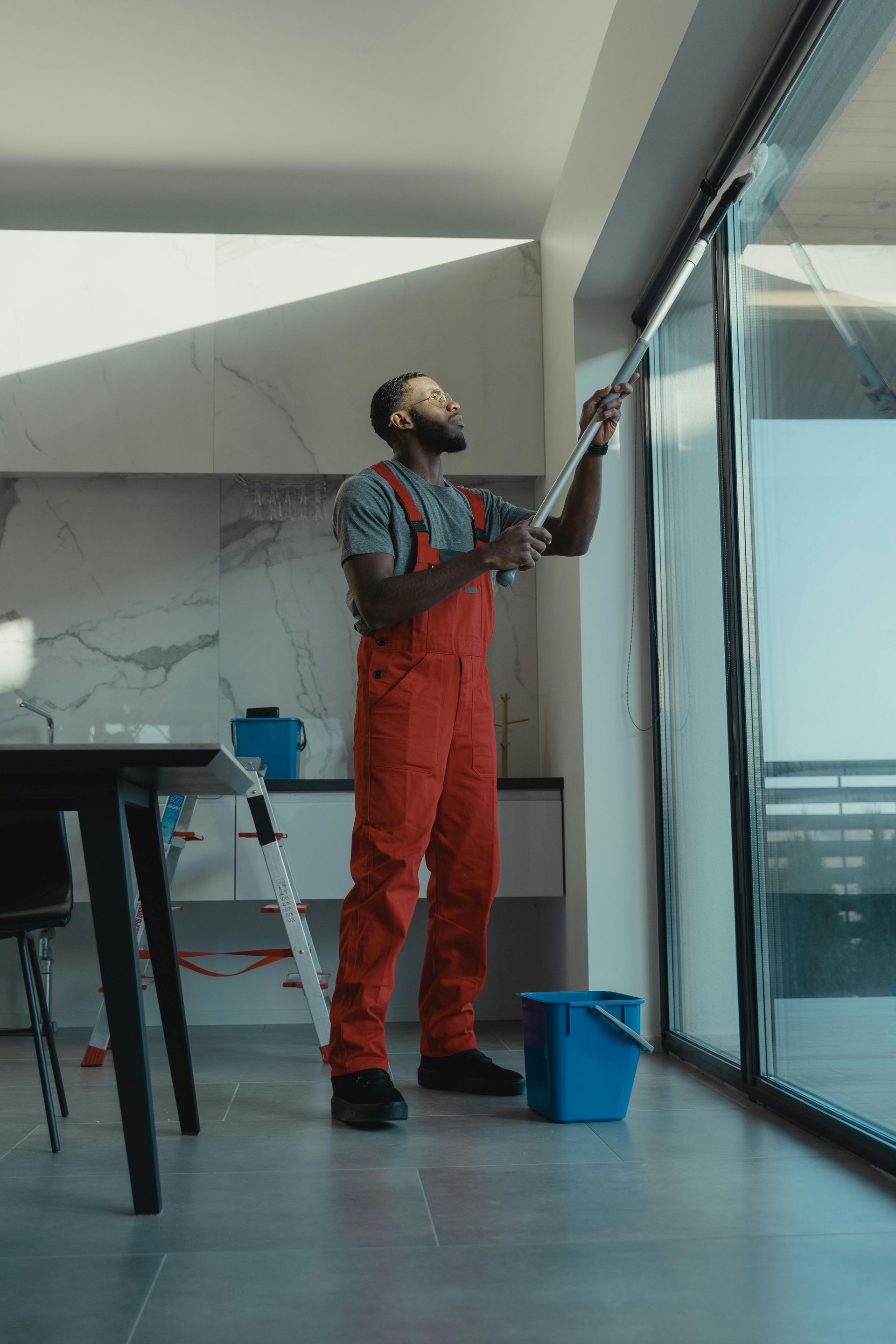 Man in red overalls cleans large window with a squeegee; a blue bucket and a ladder nearby.