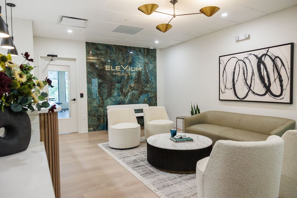 A modern waiting room featuring a marbled accent wall, two white armchairs, a tan sofa, and a round marble coffee table.