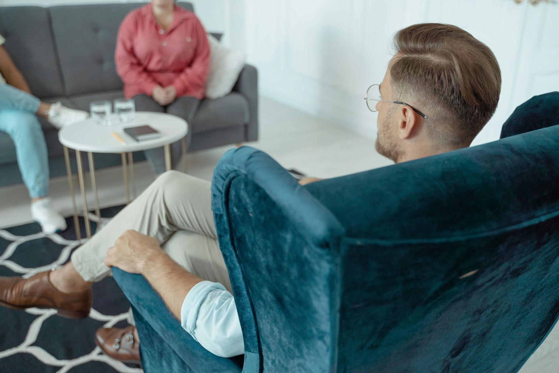 A professional seated in a teal armchair faces a client on a sofa, with a small table between them in a bright office.