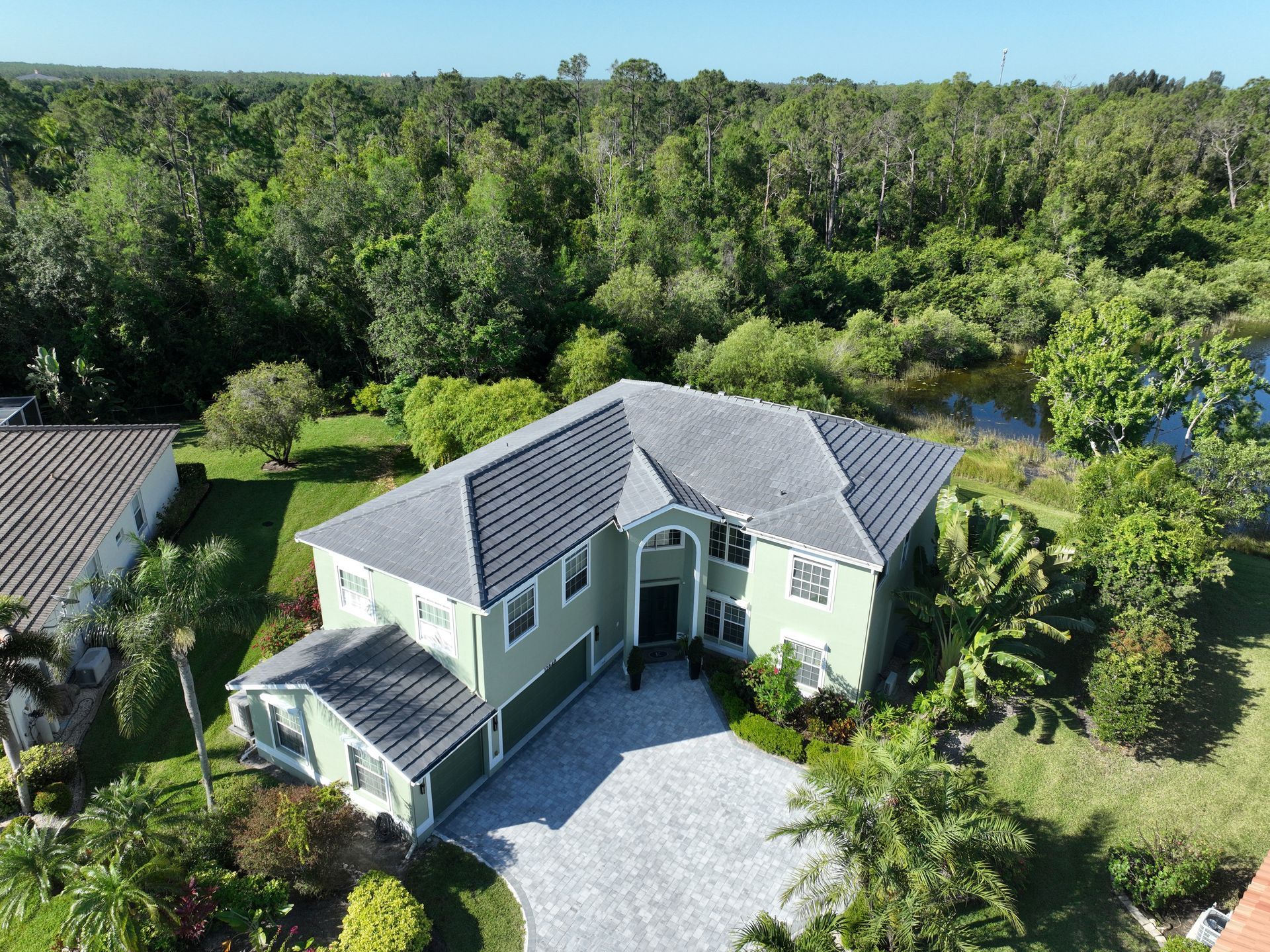 Two-story green house with gray roof and paver driveway, surrounded by greenery.