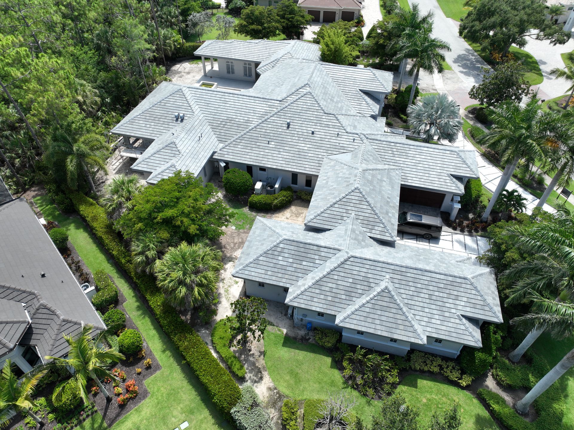 Aerial view of a large house with gray roofs, surrounded by green trees and grass, in a sunny setting.