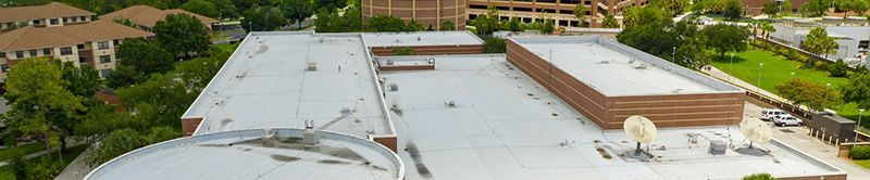 Aerial view of a building roof and surrounding green trees. White roof, with a brown brick structure and a satellite dish.