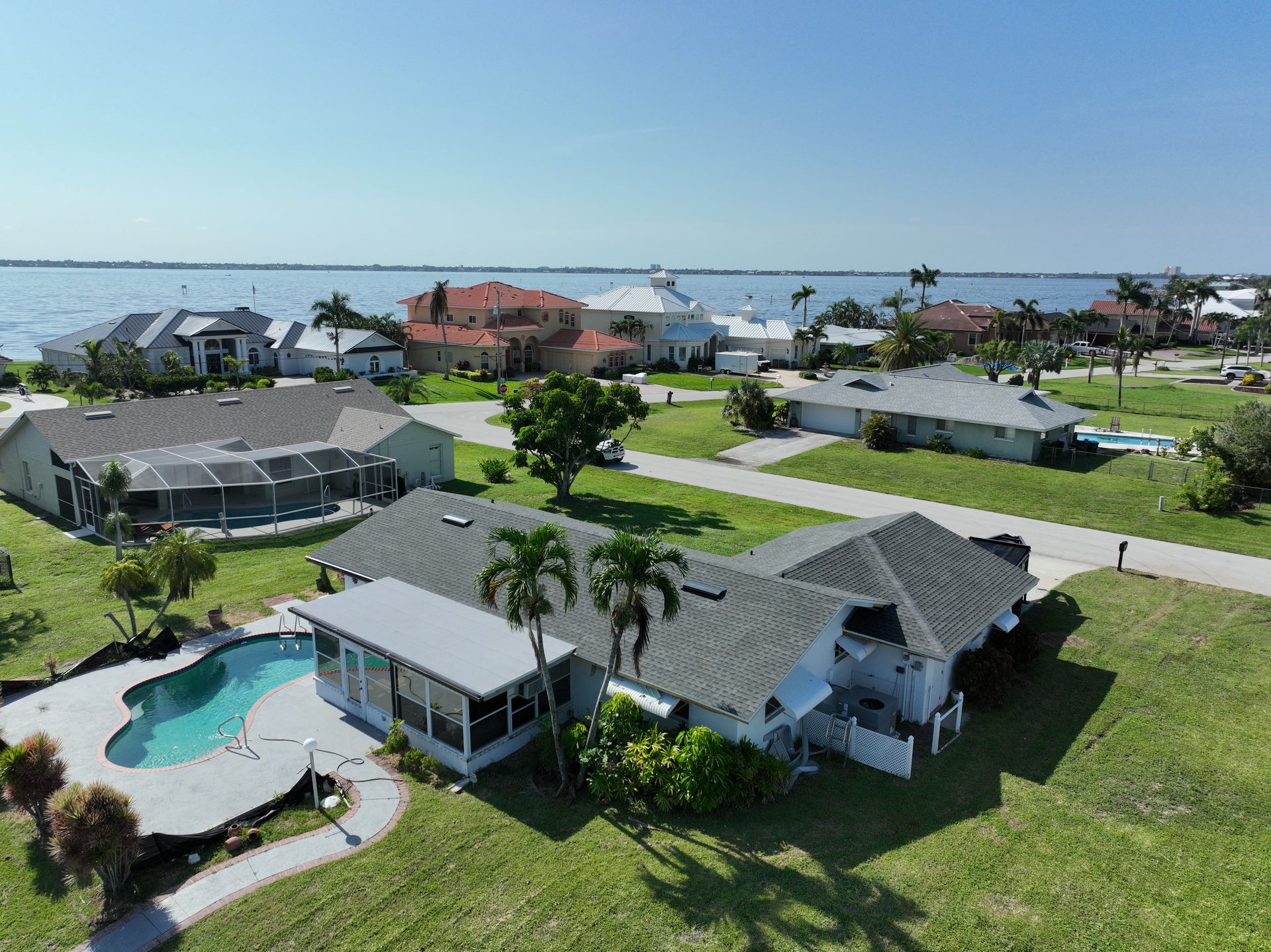 Aerial view of waterfront homes with pools, green lawns, and a blue ocean in the distance.