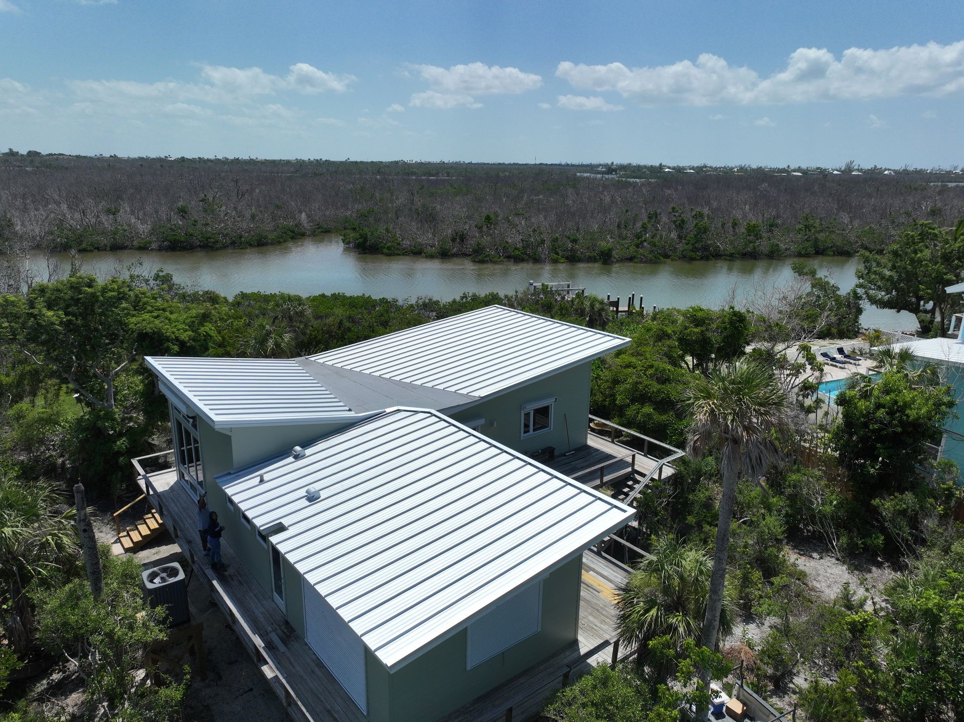 Aerial view of a light green house with a wavy silver metal roof next to water and vegetation. Blue sky.
