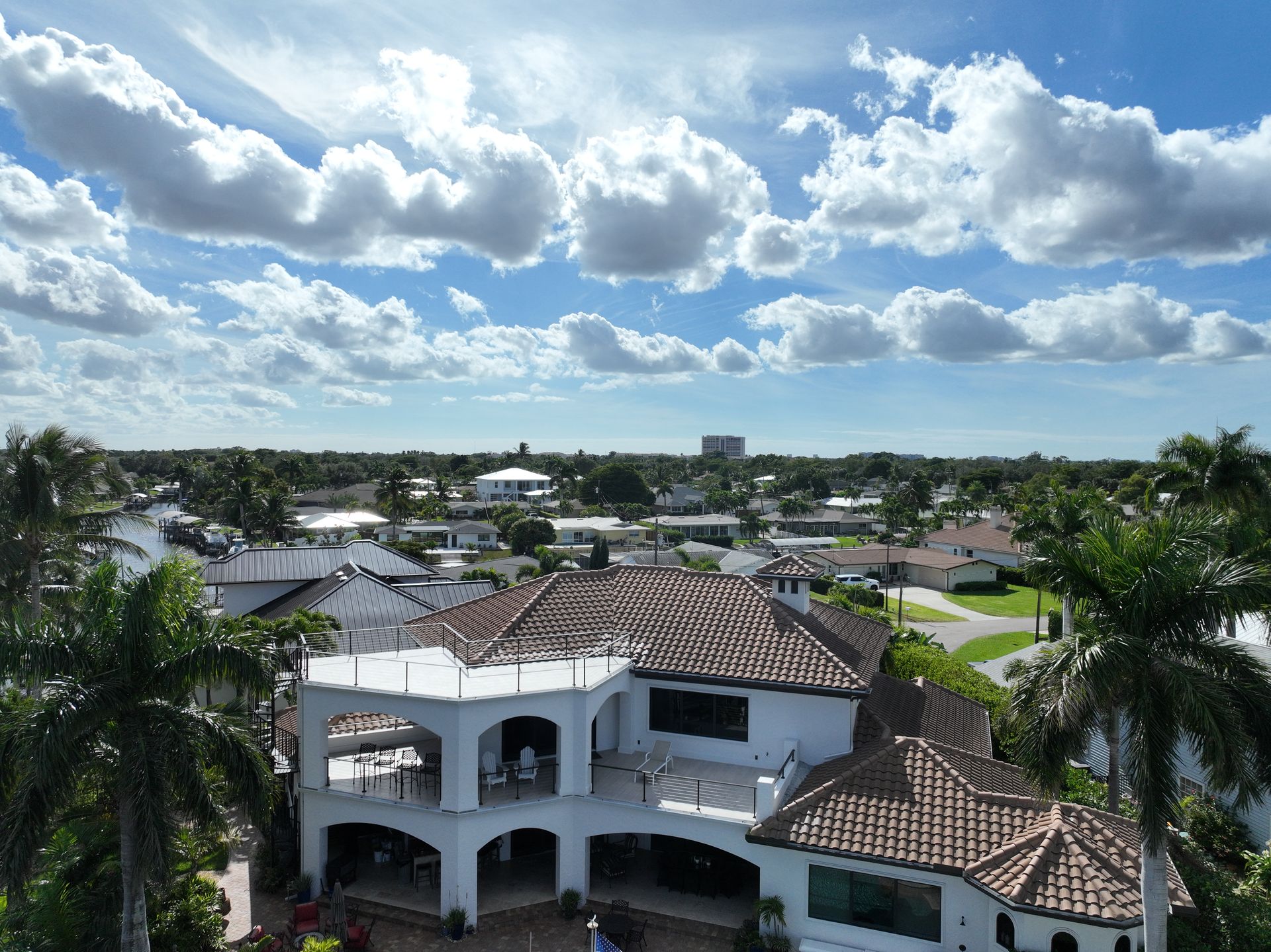 An aerial view of a large white house with a terracotta roof, palm trees, and a cloudy blue sky.