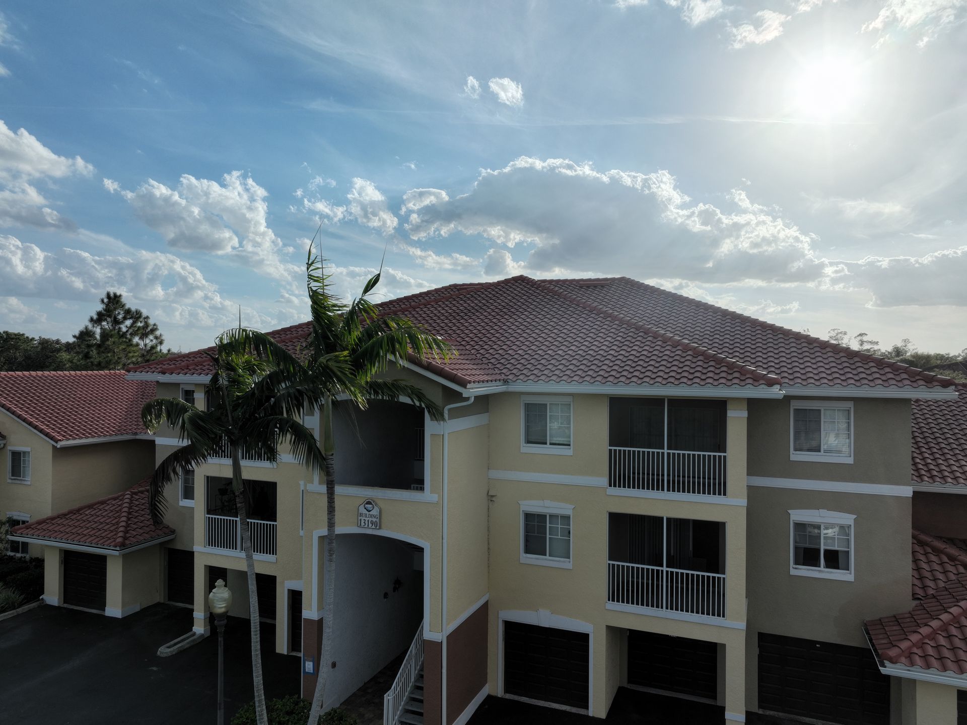 Three-story apartment building with red-tiled roof and palm trees, set against a partly cloudy sky.