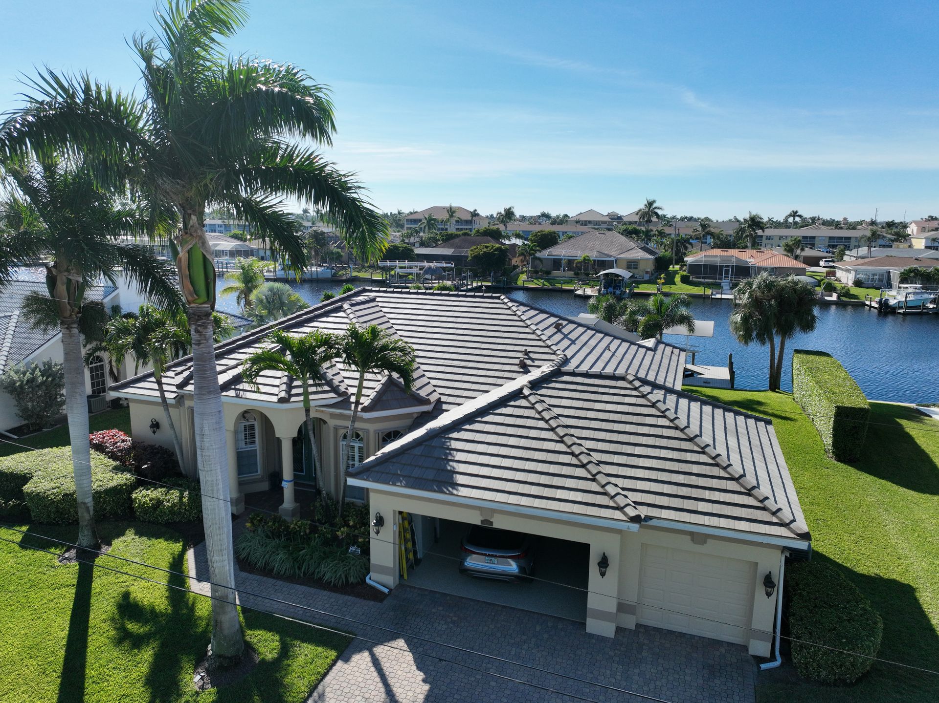 Drone view of a luxury house with a gray tile roof and a driveway, next to a canal. Palm trees are in the yard.