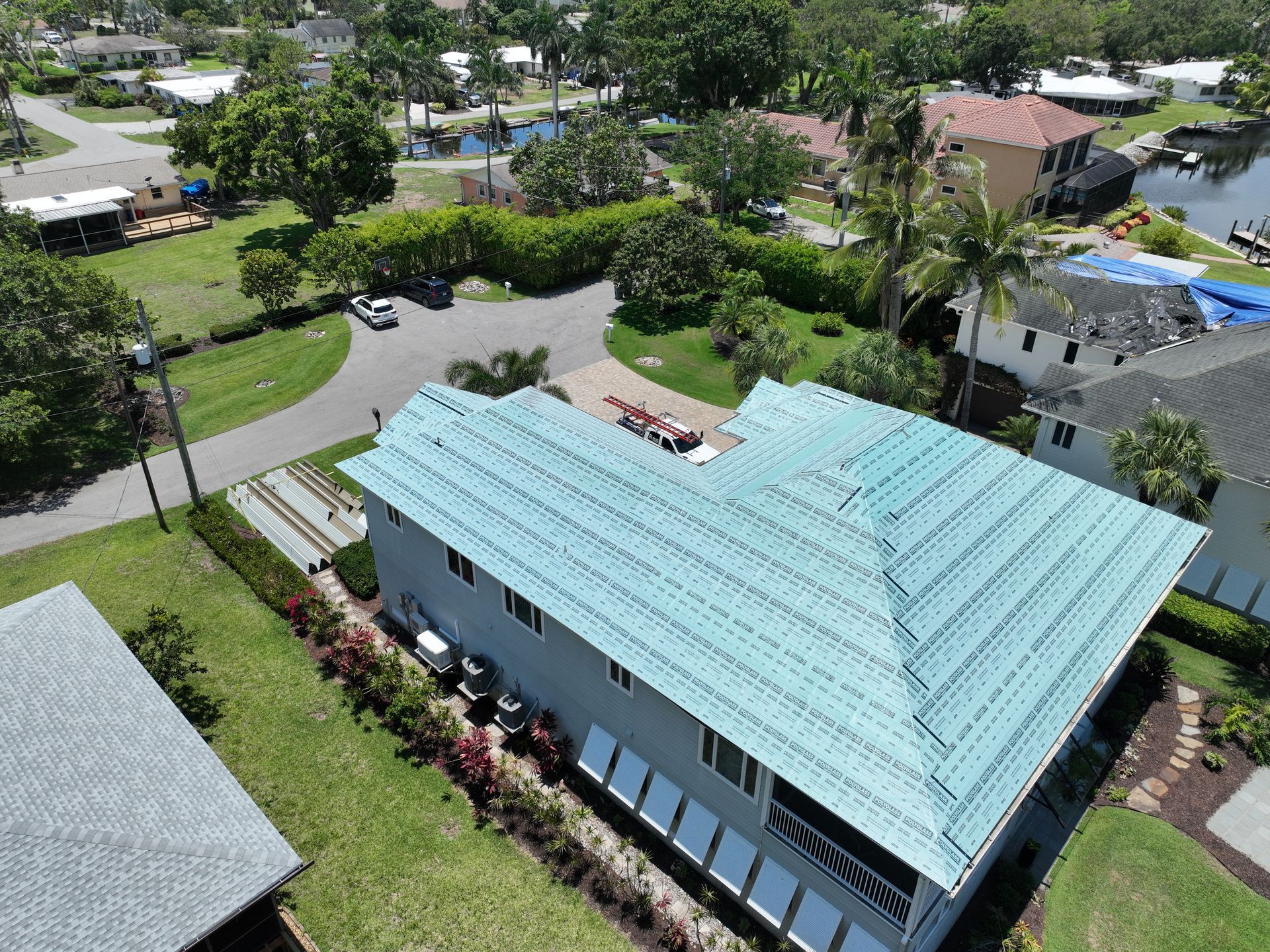 Aerial view of a two-story blue house with green roof tiles, near a canal and other homes.