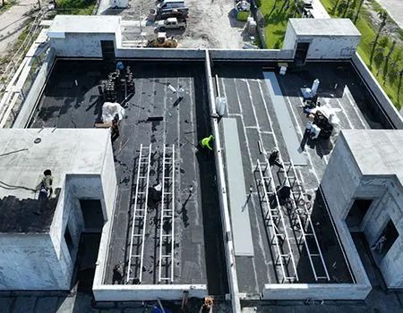 Workers installing a flat roof on a building under construction. Black tar paper, metal, and ladders are visible.