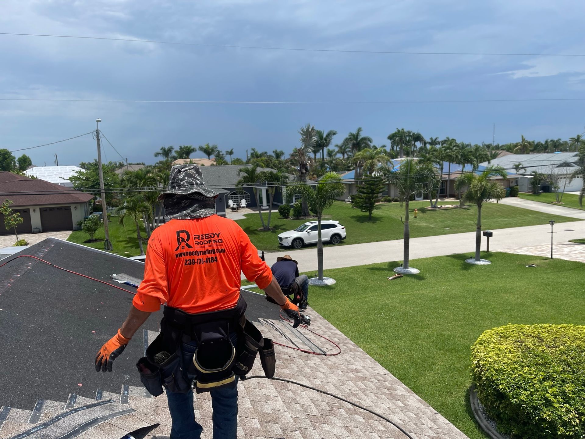 Workers on a roof in orange shirts, installing roofing materials; sunny day, residential neighborhood.