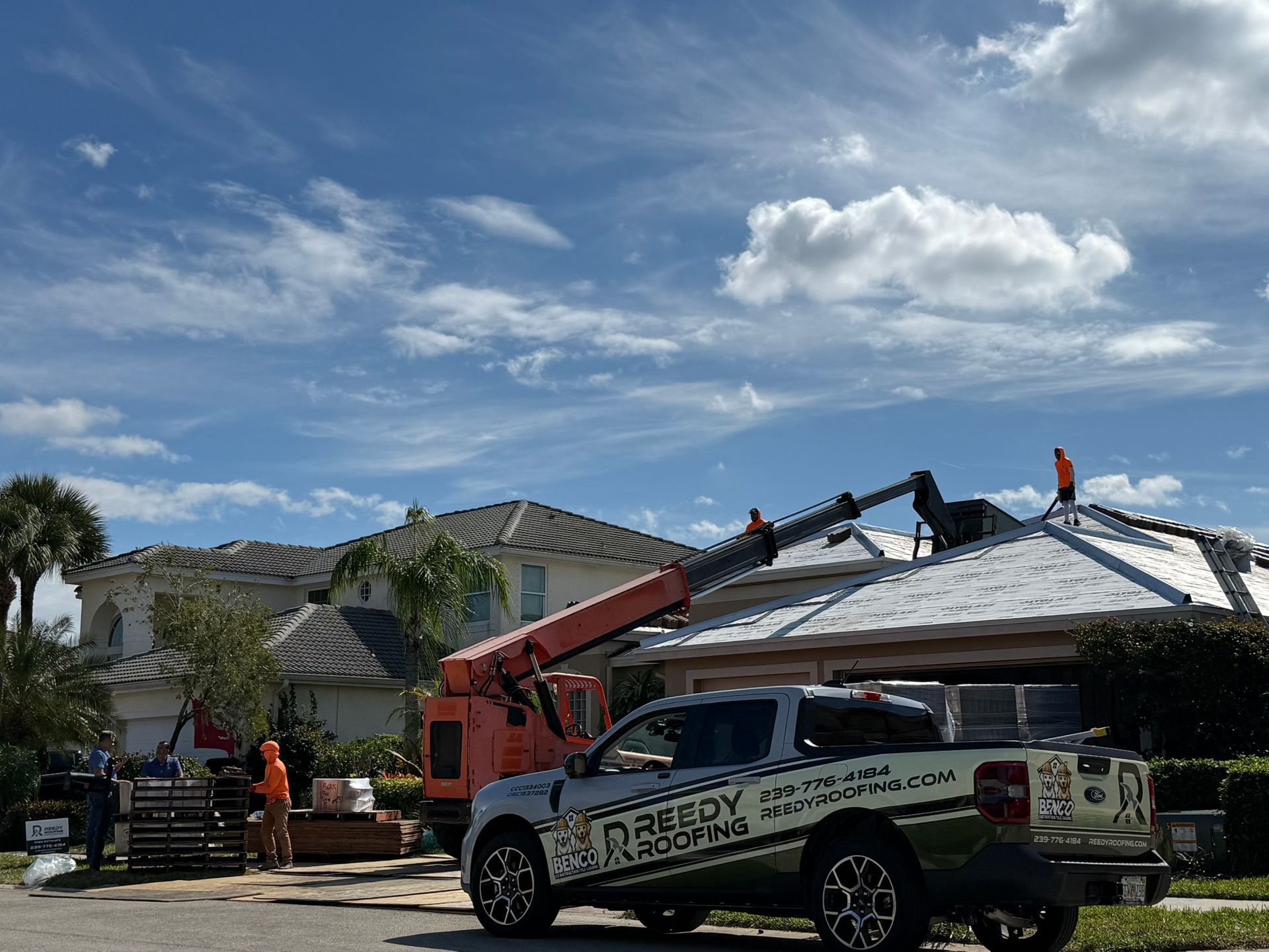 A crane assisting with roof work on a house. A work truck and workers on site under a cloudy sky.