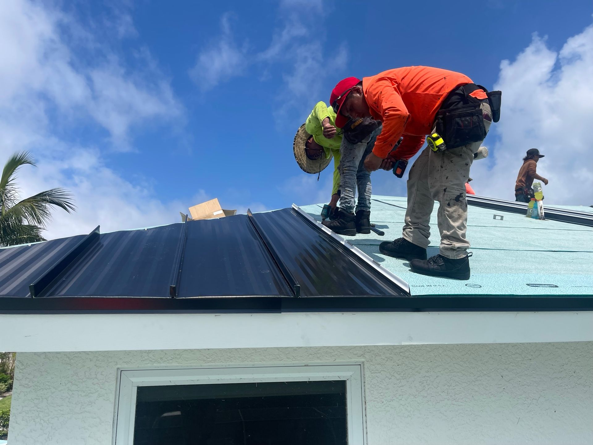 Construction workers installing black metal roof on a white house under a blue, cloudy sky.