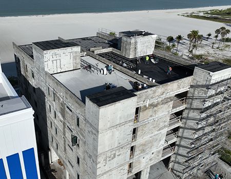 Aerial view of a concrete building under construction near a beach. Workers are on the roof.