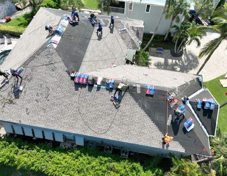 Roofing crew working on a house. Workers in blue shirts on a gray shingled roof, with materials and equipment visible.