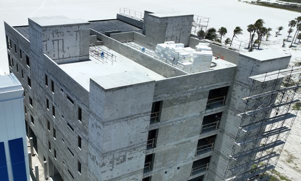 Construction of a multi-story concrete building with scaffolding and a flat roof, near palm trees and a sandy shore.