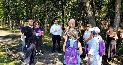 Group of people listening to a person gesturing outdoors, in a wooded area. Some wear purple.