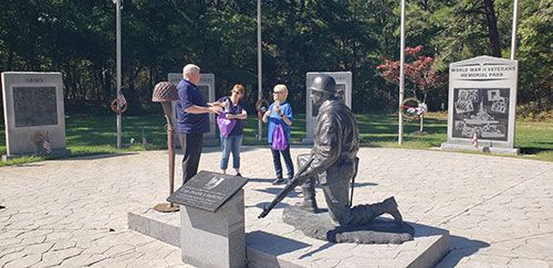 A group stands near a war memorial, including a kneeling soldier statue, plaques, and stone monuments.