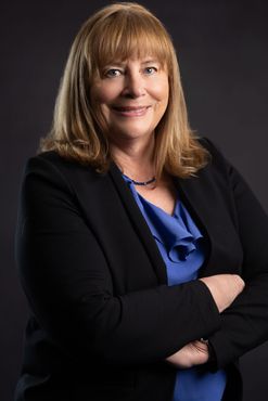 A woman in a black jacket and blue shirt is standing with her arms crossed.