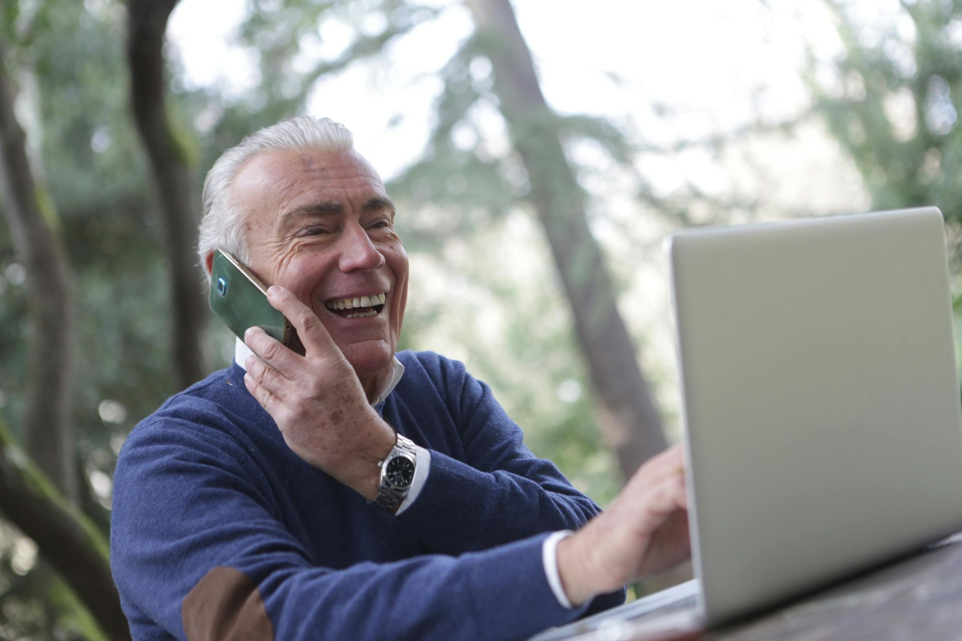 An elderly man is talking on a cell phone while using a laptop computer.