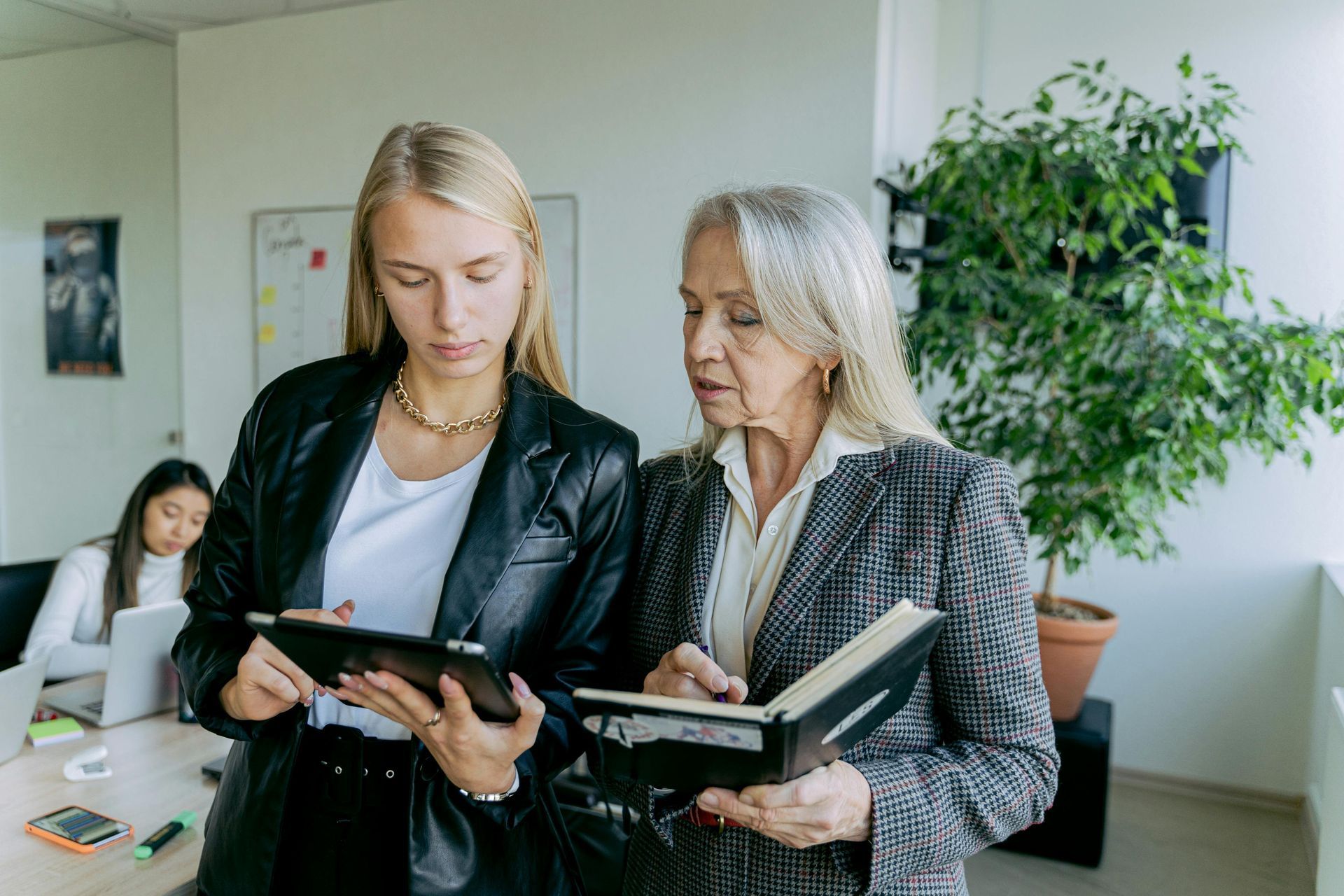 Two women are standing next to each other in an office looking at a tablet.