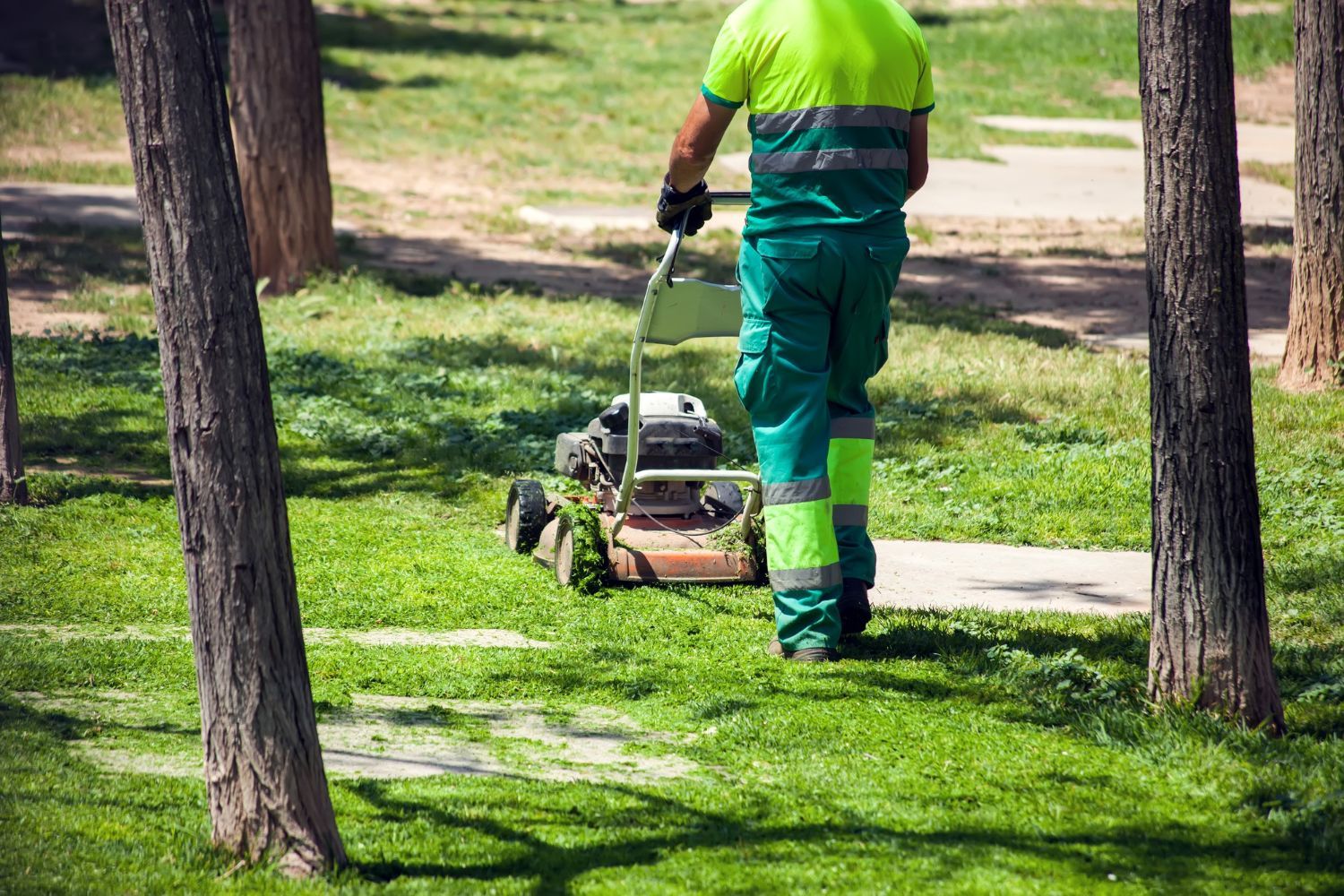 Man mowing grass in a park, wearing green work clothes and reflective stripes.