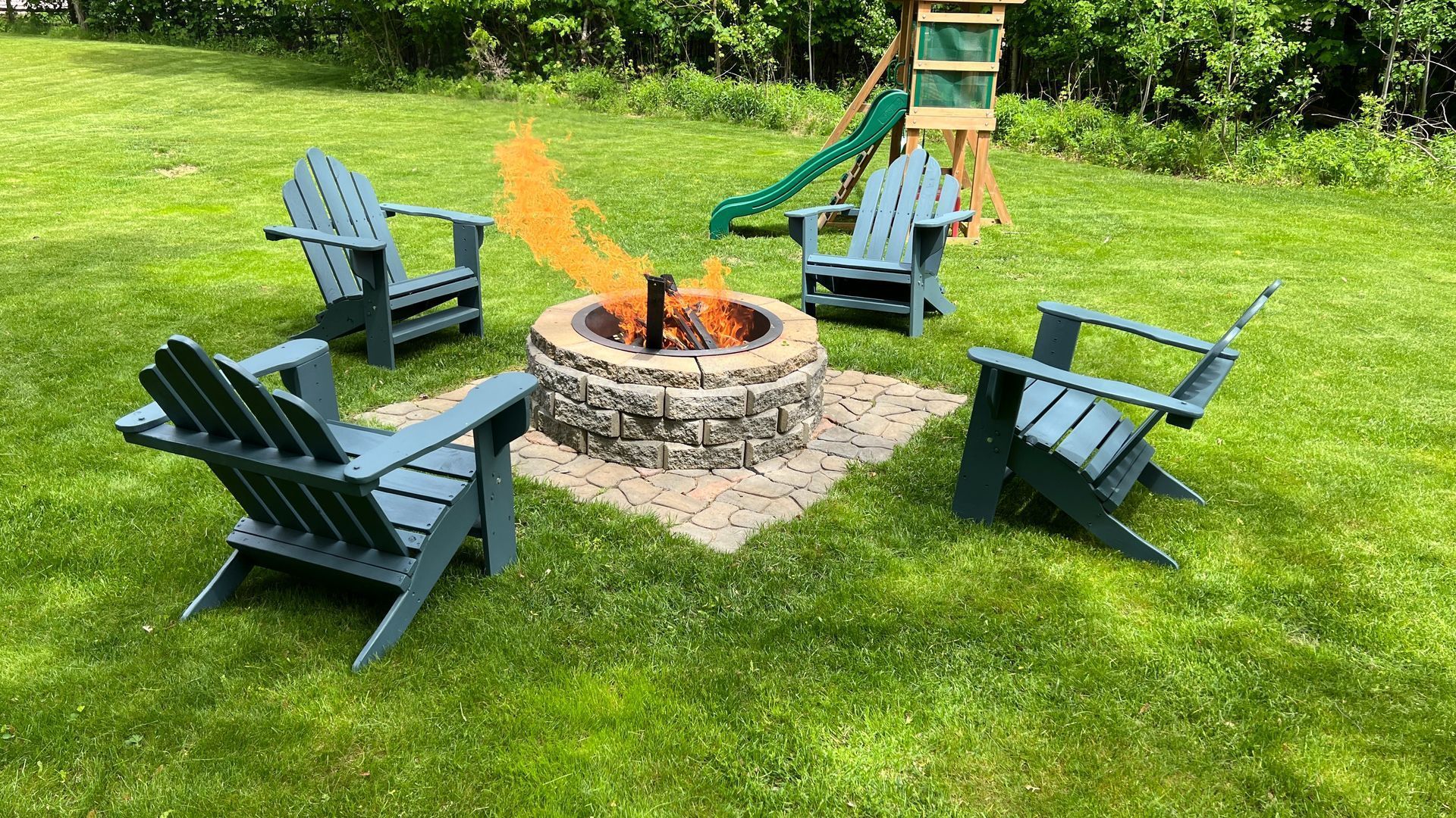 Four teal Adirondack chairs circle a lit fire pit in a grassy backyard.