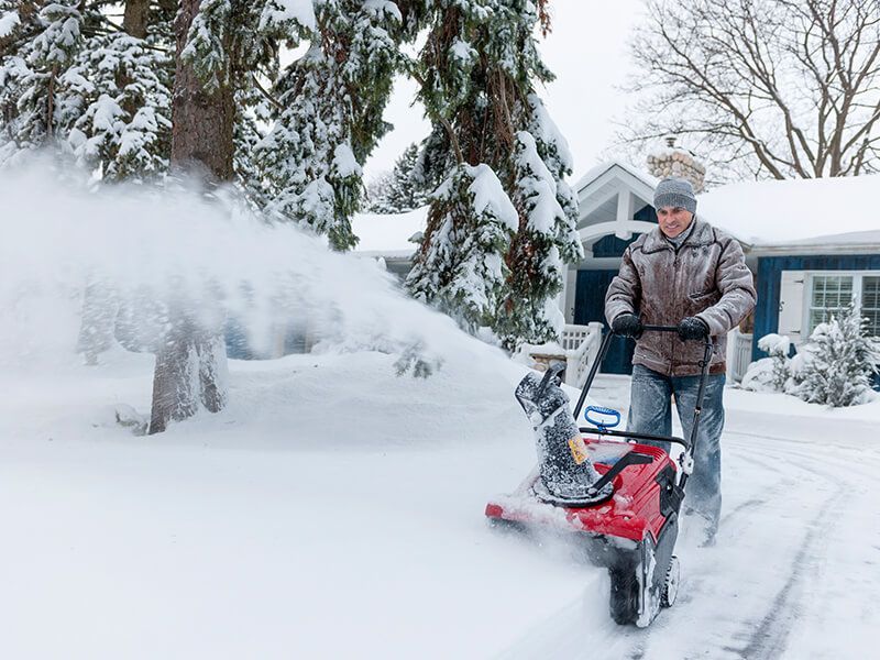Man using snowblower to clear a snowy path in front of a blue house.