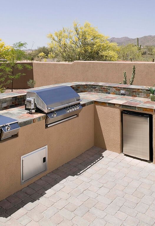 Built-in outdoor kitchen with a grill, refrigerator, and storage, set against a stucco wall and stone patio.