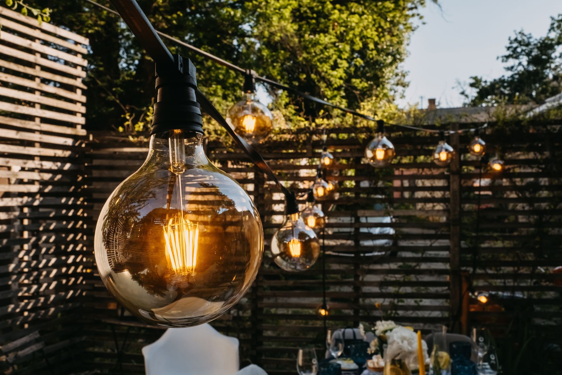 String lights with large bulbs illuminate an outdoor dining area with a wooden fence.