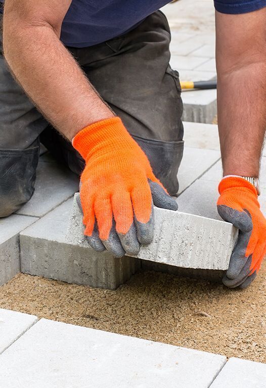 Person wearing orange gloves placing a rectangular paver, part of a patio being built.