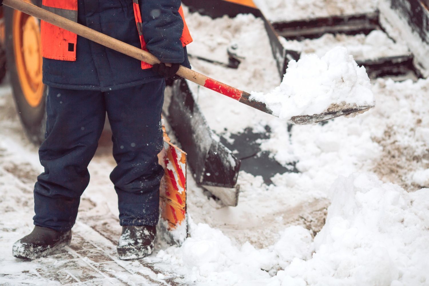 Person in blue coat and orange vest shoveling snow near a snowplow.