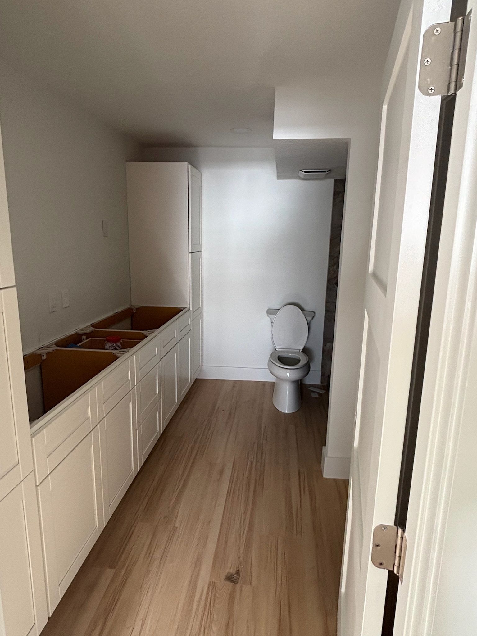 A long, narrow bathroom with white cabinets, a toilet, and light wood flooring.