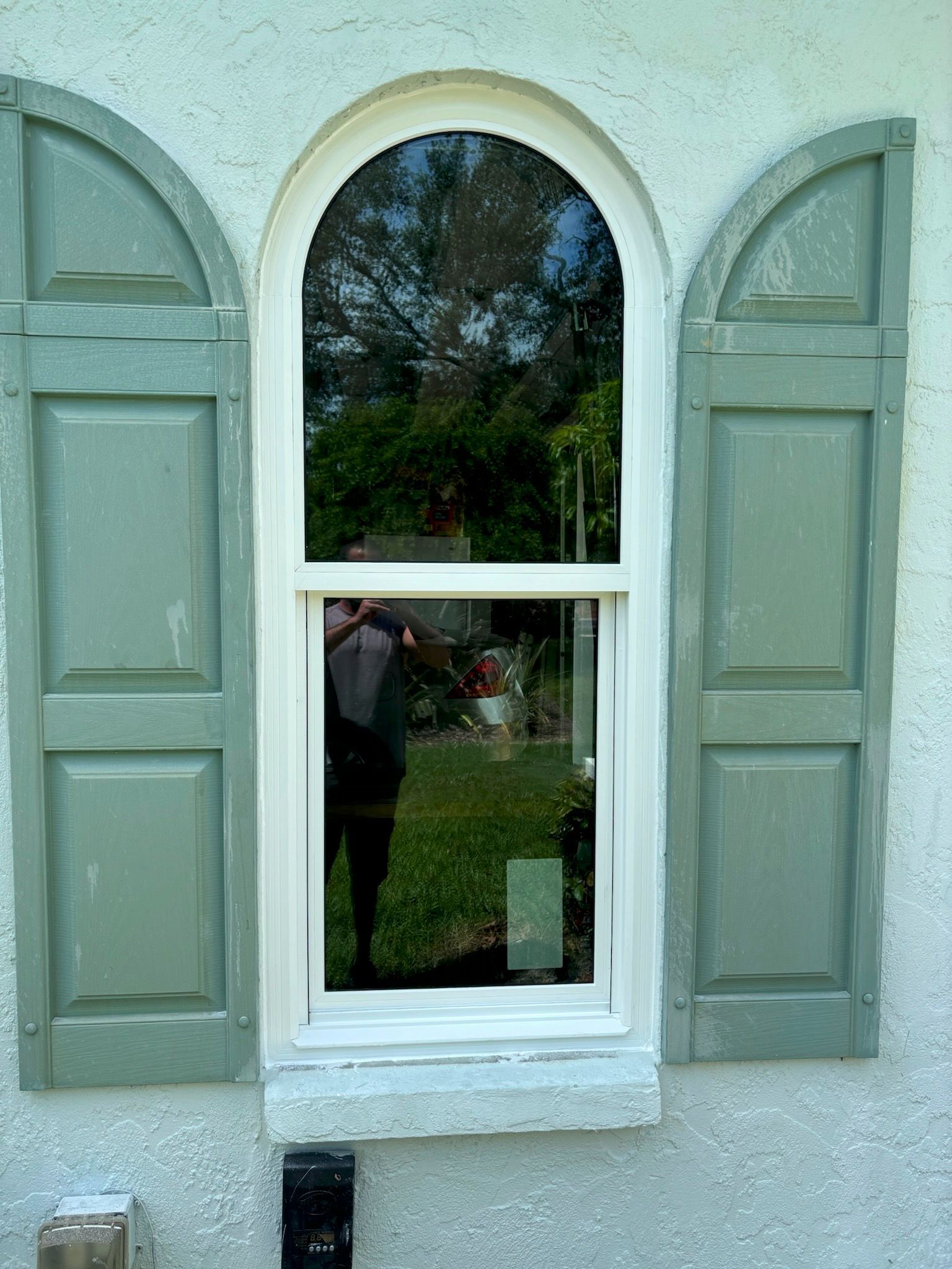 Window with green shutters, reflecting a yard, white frame, set in light blue stucco.