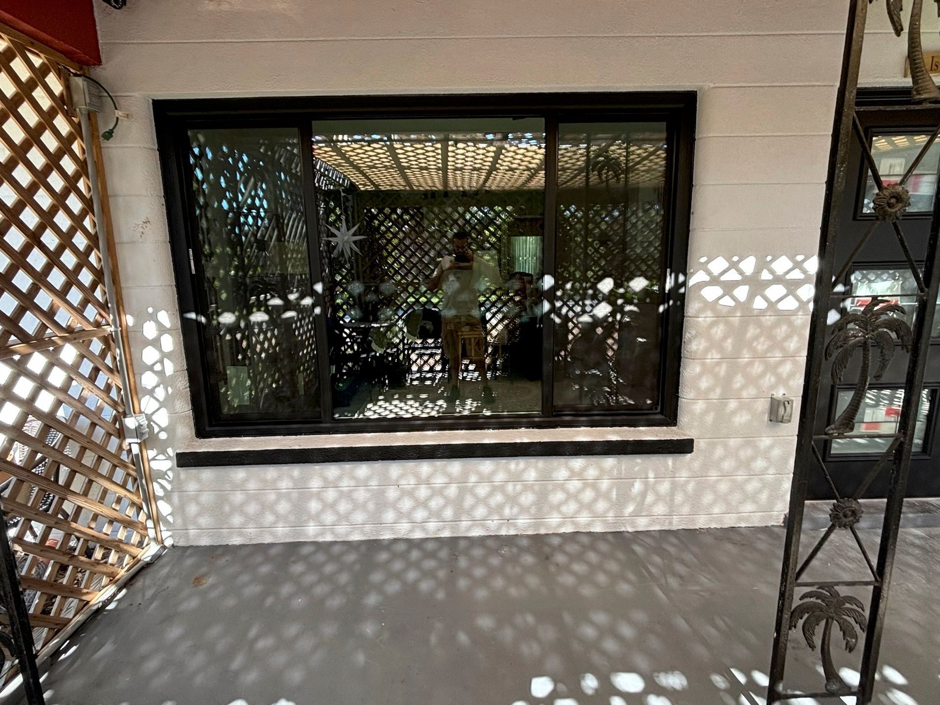 Porch with trellis, black-framed sliding window reflecting person, and gray ground. White building.
