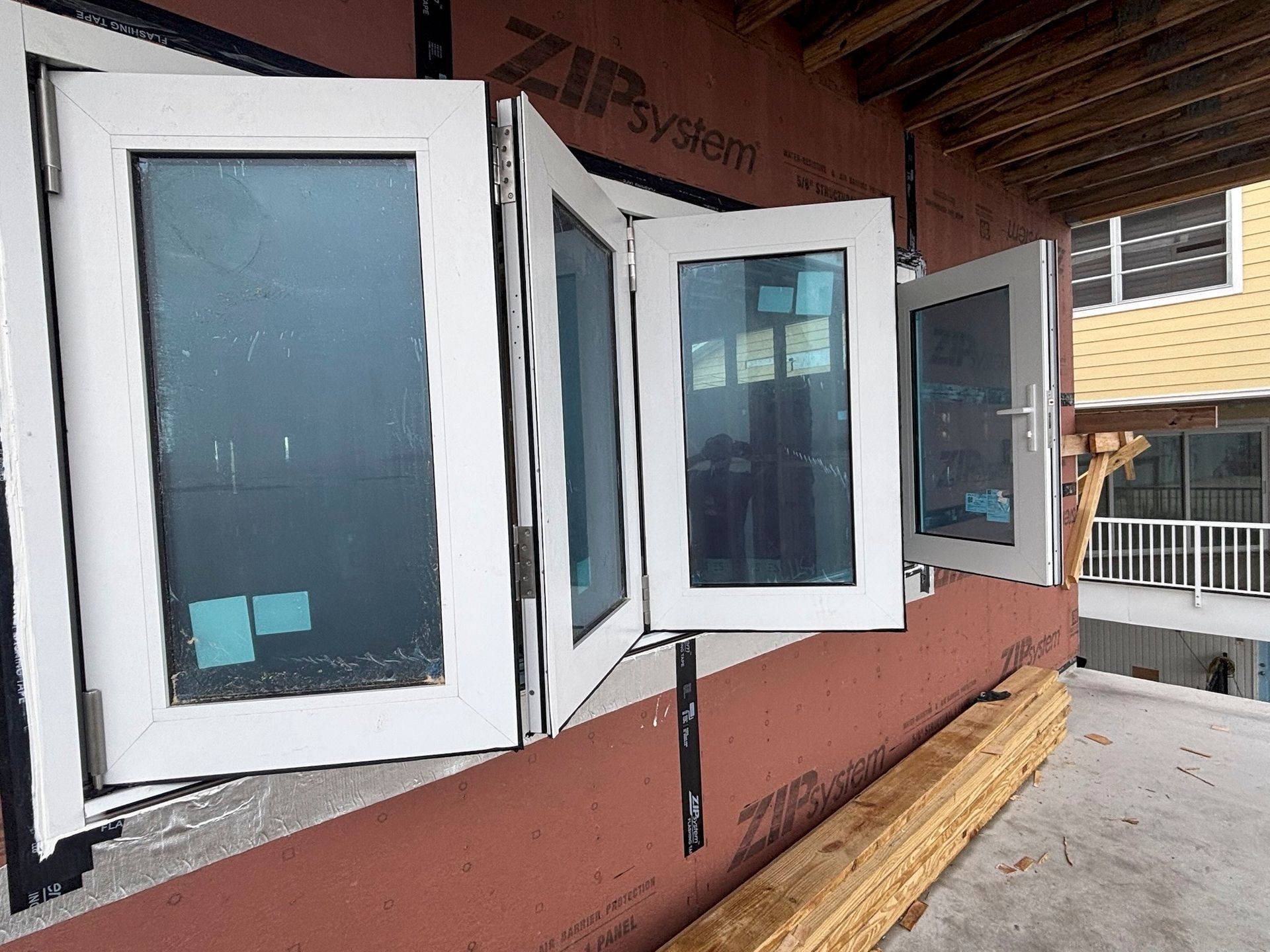 White, folding windows partially open on a building under construction, showing blue window film and a red facade.