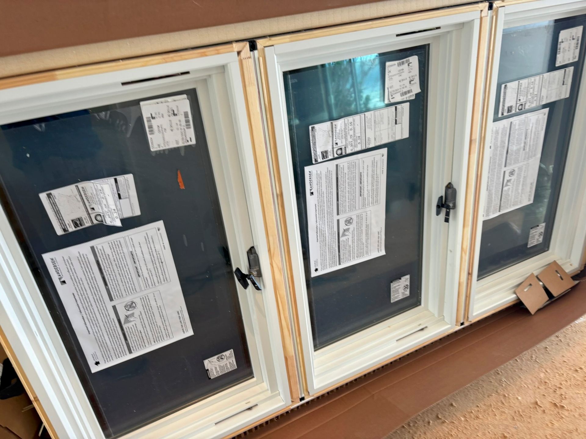 Three new white-framed windows with black sashes, covered in protective film and paperwork, sit inside a cardboard box.