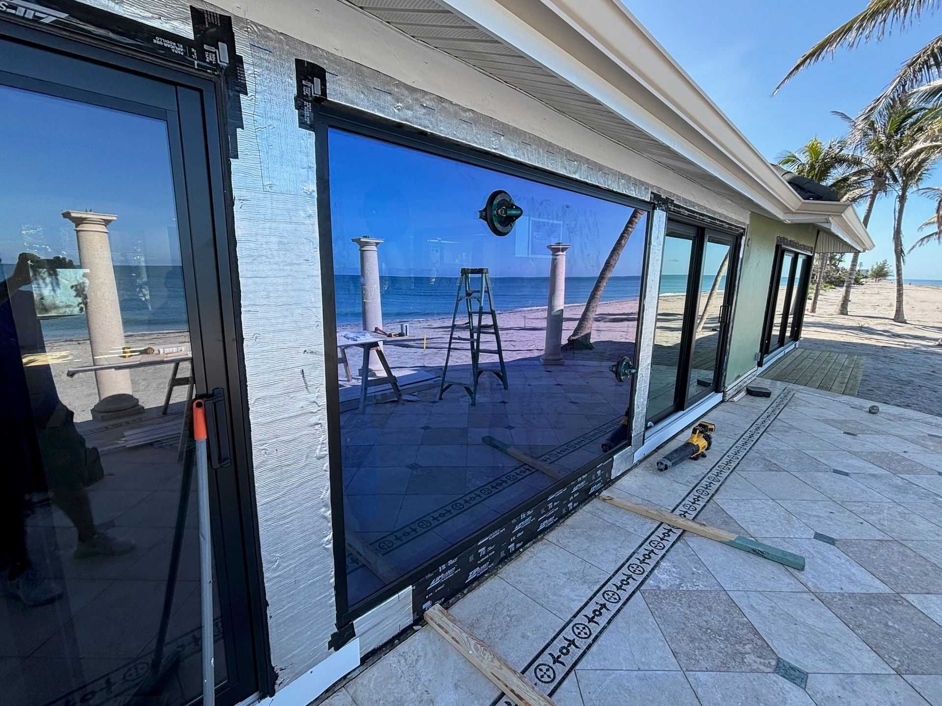 Exterior view of a house with large windows reflecting beach and sky, construction in progress.
